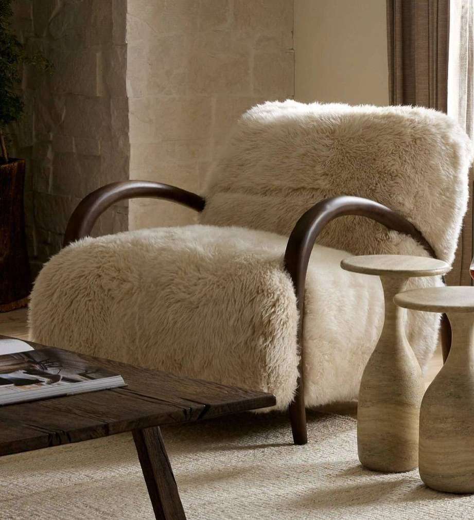 Sculptural cream shearling chair with curved dark timber arms and pale stone side tables.